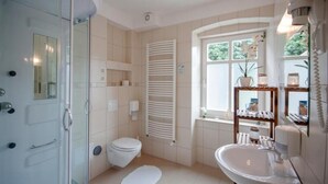 Un baño con cabina de ducha de vidrio, inodoro, lavamanos blanco y ventana con vistas a zonas verdes.