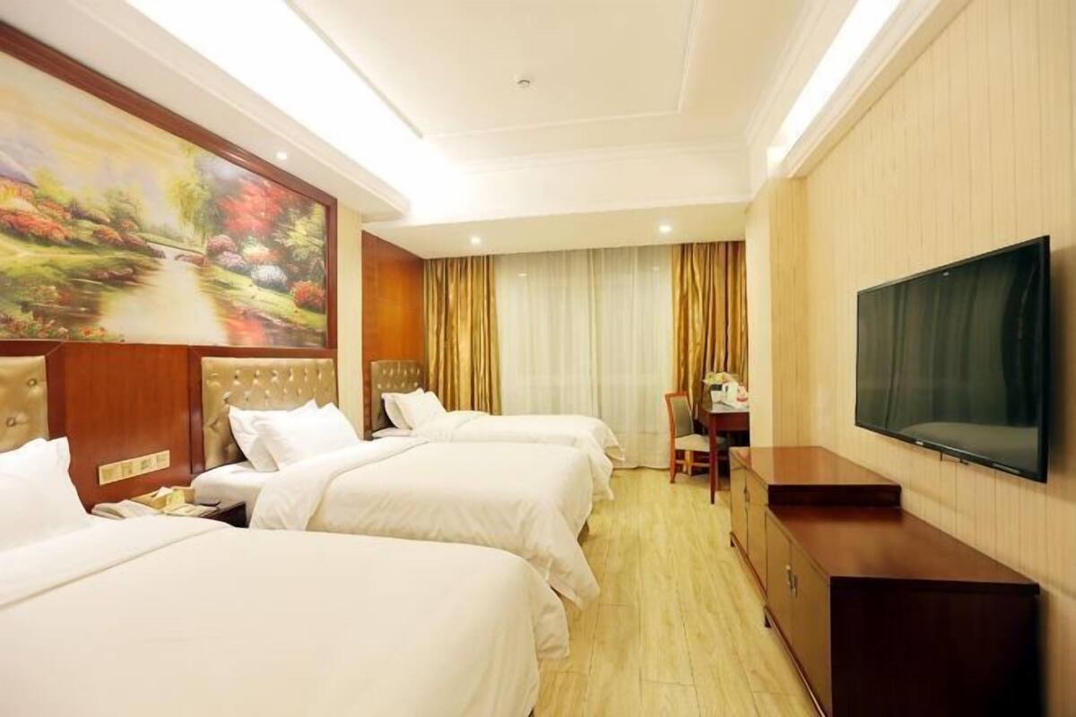 Une chambre d’hôtel avec trois lits, un bureau, une télévision et un tableau au mur.