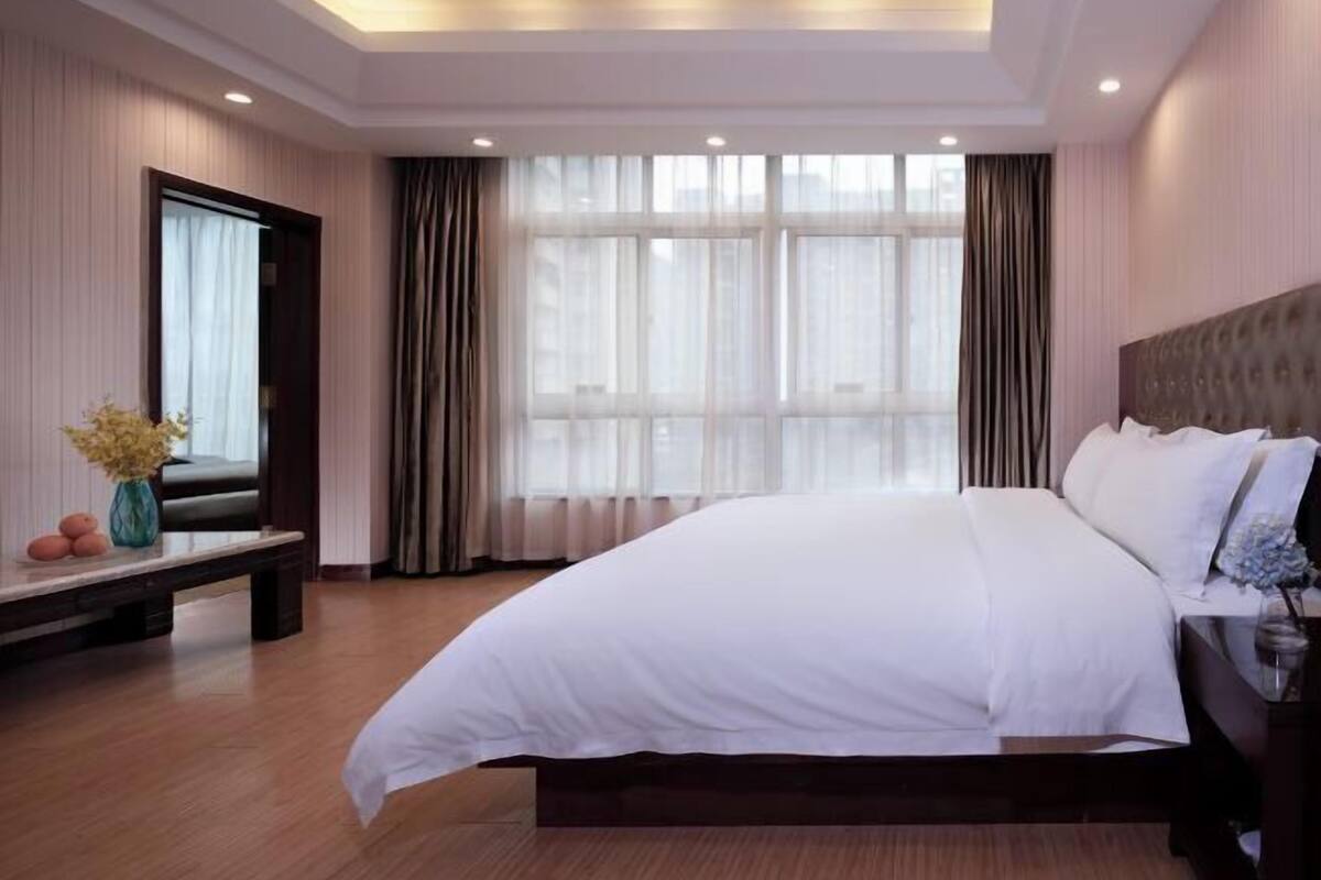 Une chambre d’hôtel avec un grand lit, une table de chevet, un banc, une télévision et une fenêtre avec des rideaux.