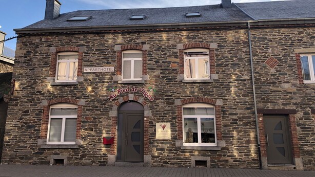 A stone building with a sign reading "Au Pied de Vigne" and "Apparhotel". It features multiple windows, a central door, and a brick facade.
