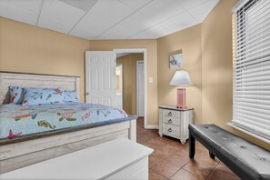 A bedroom with a bed, a nightstand, a bench, a door, and a window with blinds.