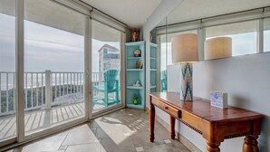 Varanda com vista para a praia e edifícios, uma mesa de madeira com abajur, e uma estante azul-turquesa com objetos decorativos.