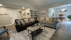 A spacious living room with a dark brown leather sofa, a grey sofa, and a glass coffee table. There is a white door and a bookshelf in the background.