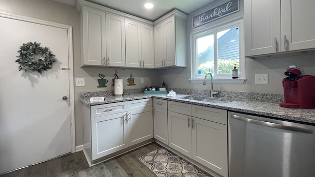 A kitchen with white cabinets, a granite countertop, a stainless steel dishwasher, and a window with a view of the outside.