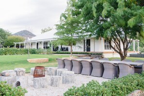 Ein Außenbereich mit Korbmöbeln und Steinpflanzgefäßen, ein weißes Gebäude mit Veranda im Hintergrund und ein Baum im Vordergrund.