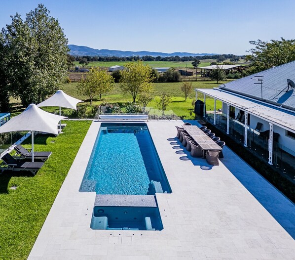 Ein rechteckiger Pool mit Whirlpool an einem Ende, umgeben von einer Terrasse mit Liegestühlen und Sonnenschirmen.