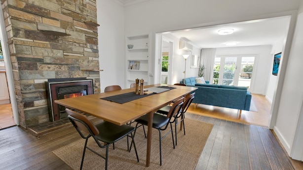 A dining area with a wooden table and chairs, a stone fireplace, and a view of a blue sofa and a balcony.