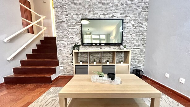 A modern living room with a wooden staircase, a flat-screen TV mounted on a textured wall, a wooden coffee table, and a built-in shelving unit with plants.