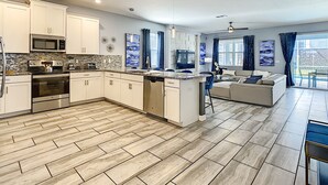 A modern kitchen with white cabinets, a microwave, stove, and dishwasher. There is a dining area with a sofa and a TV. - 8BR • Arcade & Game Room • 2 Theaters • Pool & Spa (Davenport)