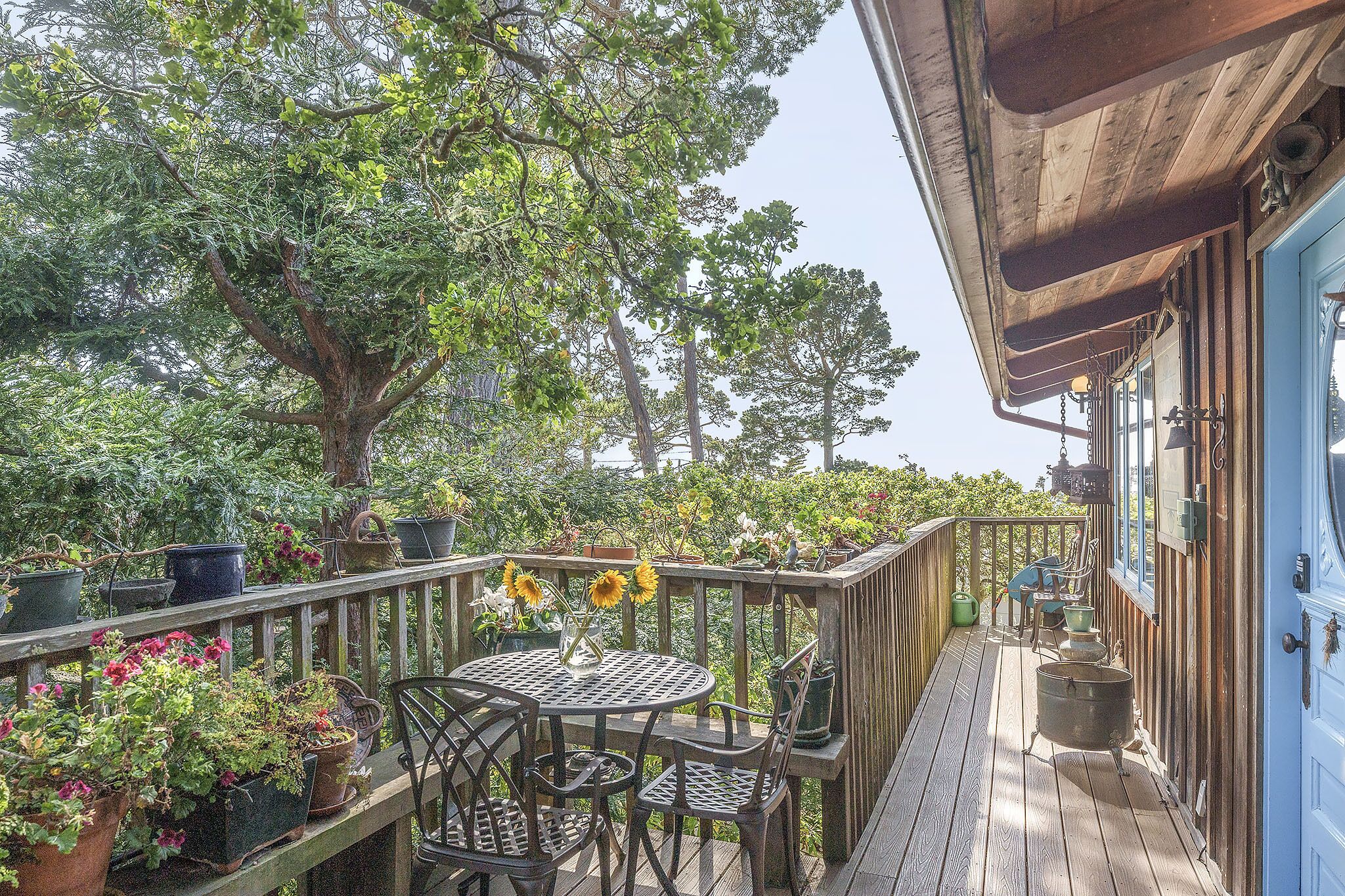 Une terrasse en bois avec une table ronde et des chaises, entourée de plantes et de fleurs en pot.