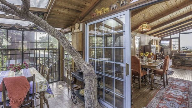 A dining area with a large tree trunk centerpiece, a glass door leading to a kitchen, and a view of the outdoors. - 3879 Lucy's Treehouse ~Unique Vintage Redwood Home (Carmel)