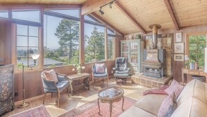 A cozy room with wooden beams, large windows, a fireplace, and comfortable seating. - 3879 Lucy's Treehouse ~Unique Vintage Redwood Home (Carmel)