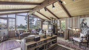 A spacious living room with a high ceiling, large windows, and a cozy fireplace. - 3879 Lucy's Treehouse ~Unique Vintage Redwood Home (Carmel)
