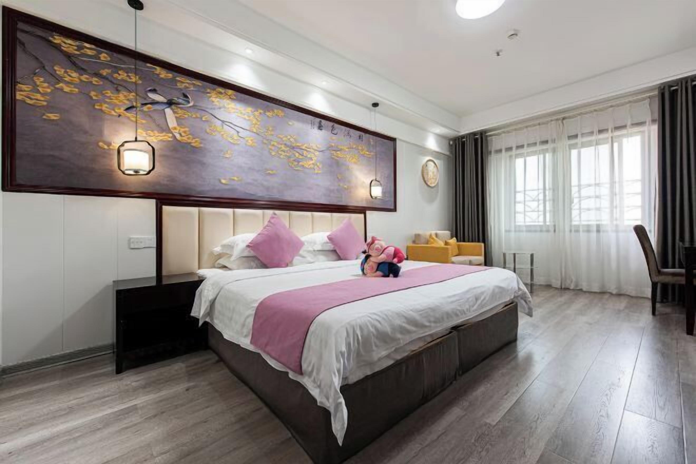 A modern hotel room with a large bed, a headboard with integrated lighting, a decorative wall piece, and a window with curtains.
