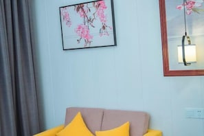 A room with a light blue wall, a framed floral artwork, a mirror with a light fixture, and a sofa with yellow and pink cushions. - Xingtian Hotel (Yueyang)