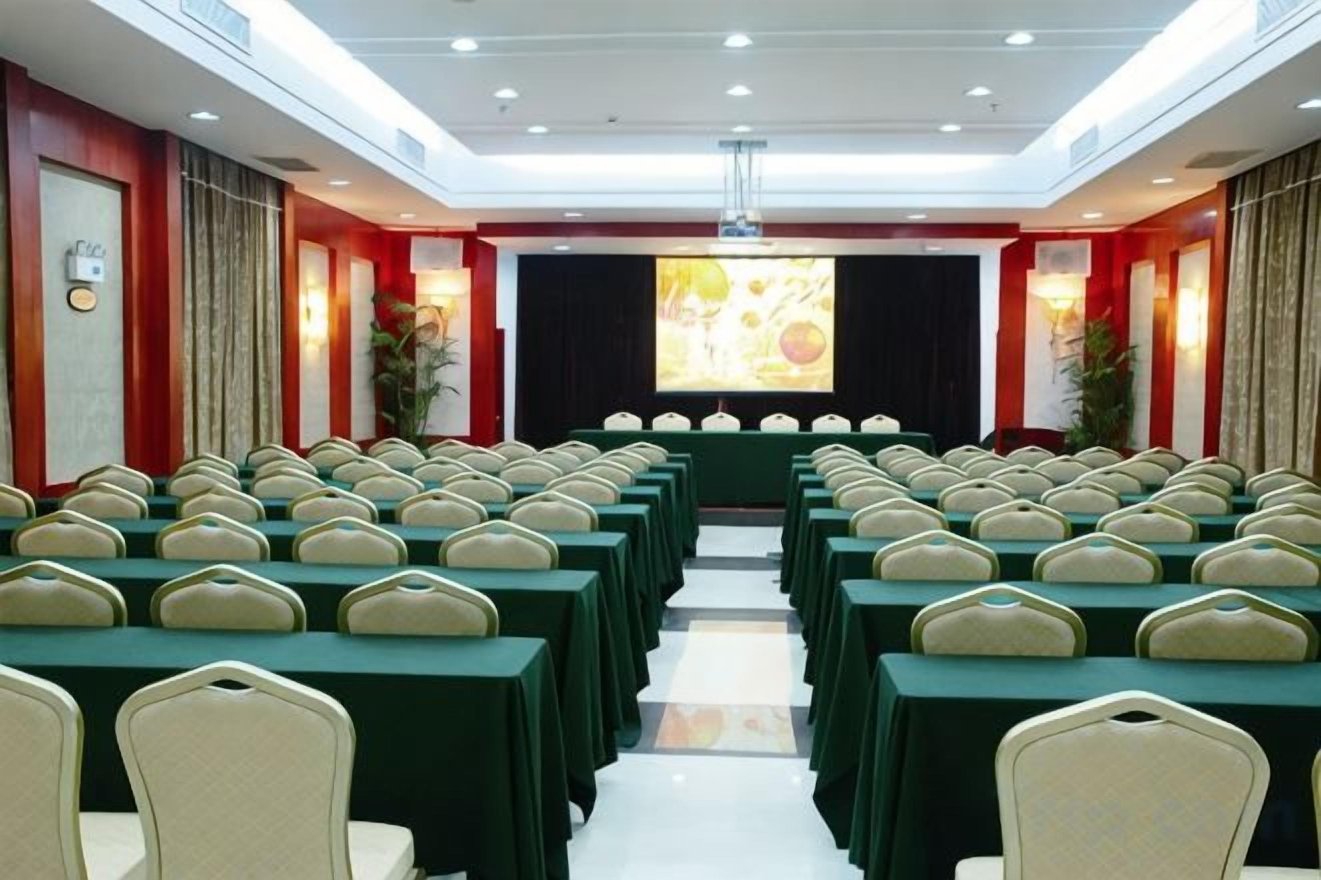 A conference room with rows of tables and chairs, a stage with a projector screen, and a red accent wall.