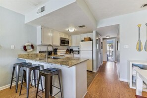 A modern kitchen with white cabinets, a granite countertop, and stainless steel appliances. There are three bar stools and a small island with a sink.