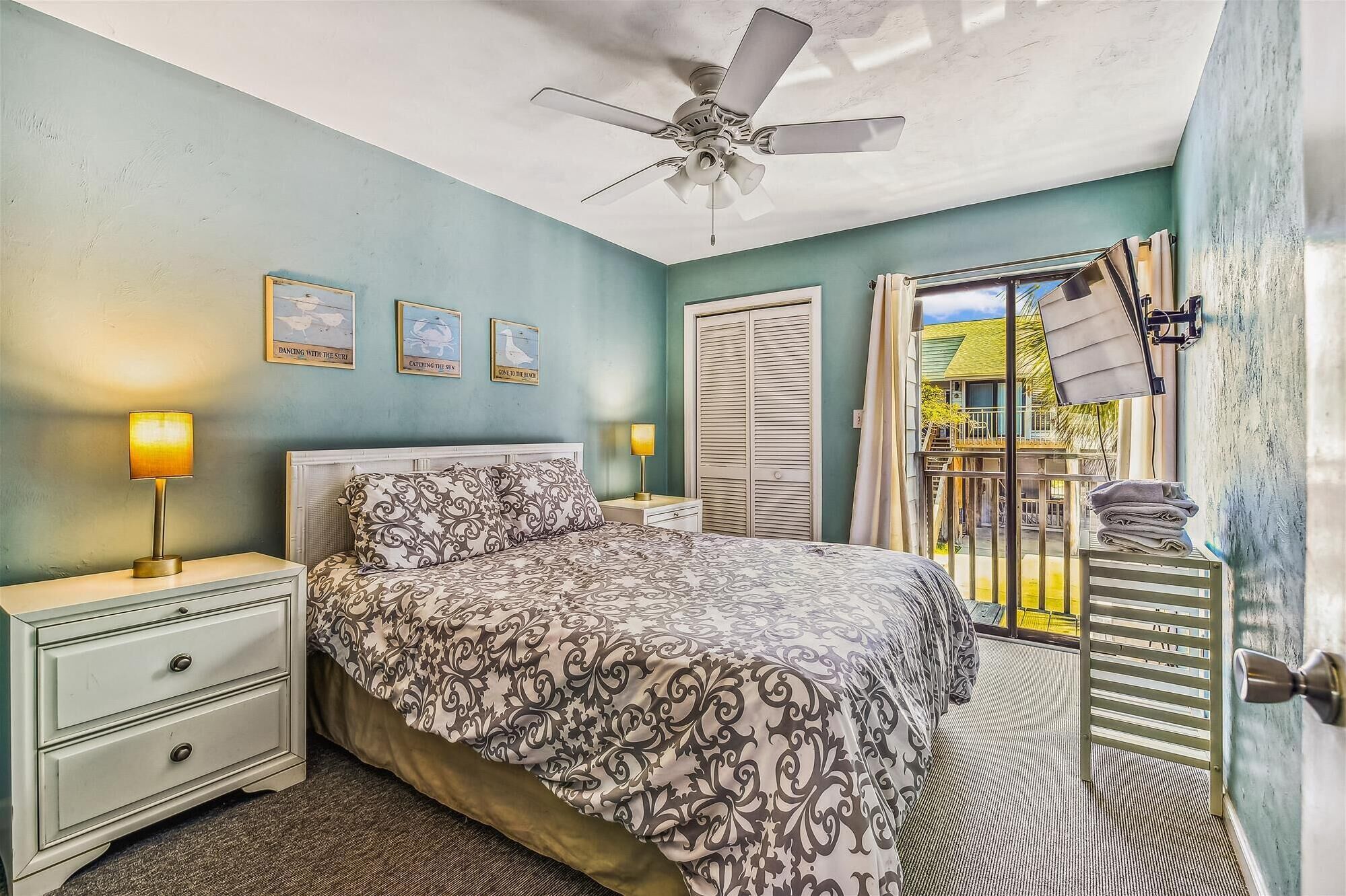 A bedroom with a bed, bedside lamps, a ceiling fan, and a view of the outside.