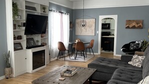 A modern living room with a flat-screen TV, a dining area with a table and chairs, a gray sofa, and a ceiling fan. - Updated kitchen, Close to downtown, Pet friendly, Privacy fenced backyard! (Kearney)