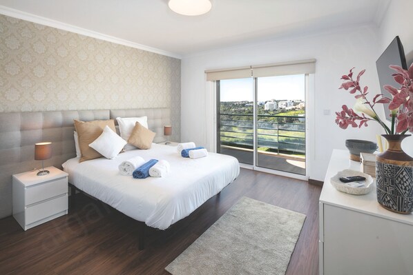 A bedroom with a large bed, a nightstand, a desk with a vase, and a view of a balcony.