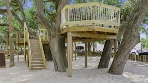 Eine Holz-Baumhauskonstruktion mit Treppe und Plattform, umgeben von Bäumen und sandigem Boden.