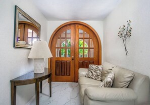 A room with a wooden door, a mirror, a table with a lamp, a sofa with patterned cushions, and a wall-mounted plant.