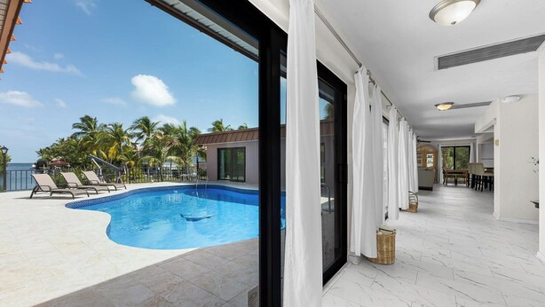 A modern hotel room with a large window overlooking a swimming pool and palm trees, a dining area, and a kitchen. - Blue Water is Calling! 5bedroom 3bath with large pool and 150 ft of dockage 2 kayaks included (Marathon)