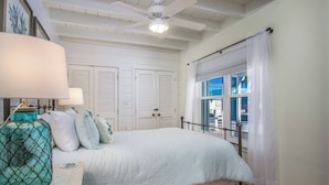 Um quarto com cama, mesa de cabeceira, janela com cortinas e ventilador de teto.