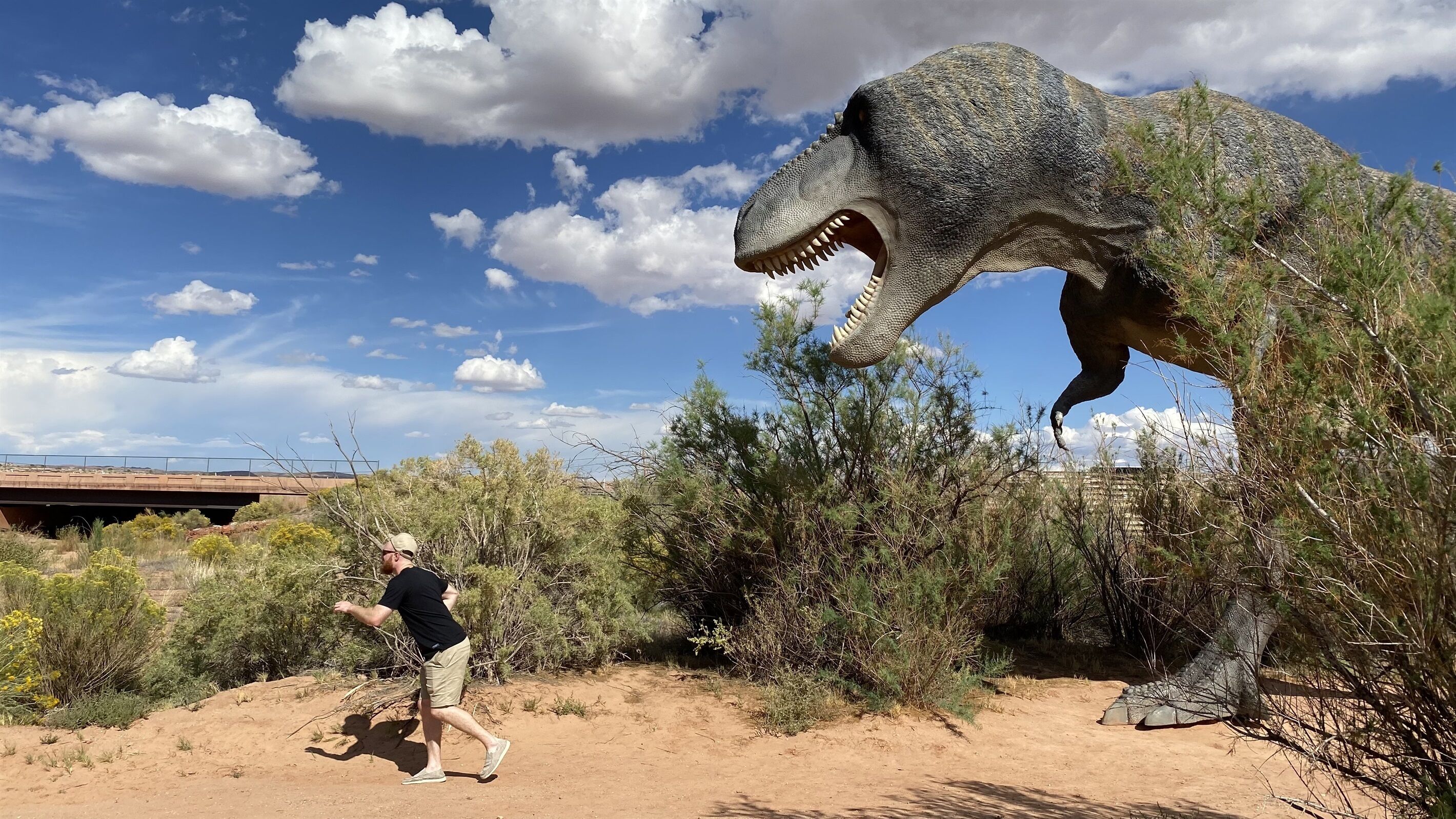 A person running towards a large dinosaur statue in a desert landscape with sparse vegetation and a clear sky.