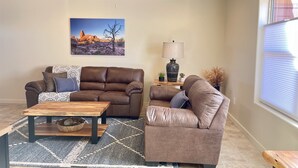 Ein Wohnzimmer mit zwei braunen Ledersofas, einem Holztisch, einem gemusterten Teppich, einer Wandlampe und einem gerahmten Bild von einer Wüstenlandschaft.