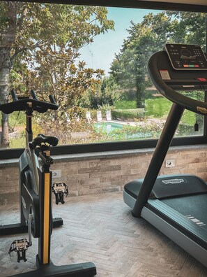 Ein Laufband und ein Ergometer in einem Raum mit großem Fenster mit Blick auf einen Garten und ein Schwimmbad.