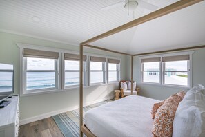 Un dormitorio con cama con dosel, ventanas amplias con vistas al océano, un escritorio con silla y suelo de madera.