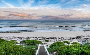 Una playa con una escalera que baja a la orilla, rodeada de vegetación.