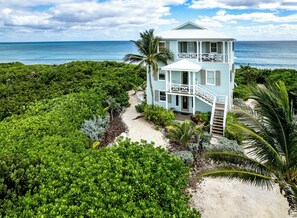 A two-story light blue house with a balcony, surrounded by lush greenery and a sandy path leading to the entrance.
