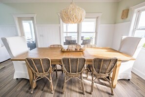 A dining area with a wooden table and wicker chairs, large windows with white trim, and a hanging light fixture.