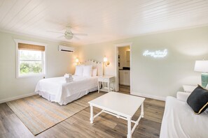 A hotel room with a bed, bedside table, a small table, a ceiling fan, and a neon sign on the wall.