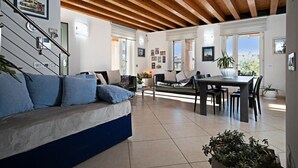 Amplia sala de estar con sofá, mesa de comedor y sillas. Se observa una escalera con barandilla, y ventanas grandes que permiten la entrada de luz natural.