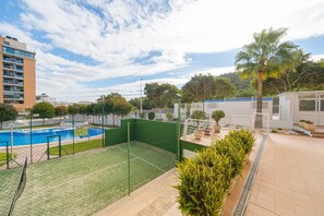 A tennis court with a pool, surrounded by greenery and a building.