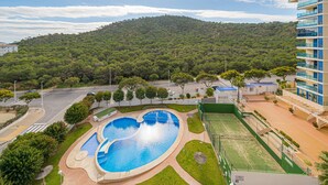 A swimming pool with a heart-shaped design, surrounded by a landscaped area with a tennis court and a building with balconies. - Apartment 'Beach and Green' with Sea View, Wi-Fi and Air Conditioning (Alicante)