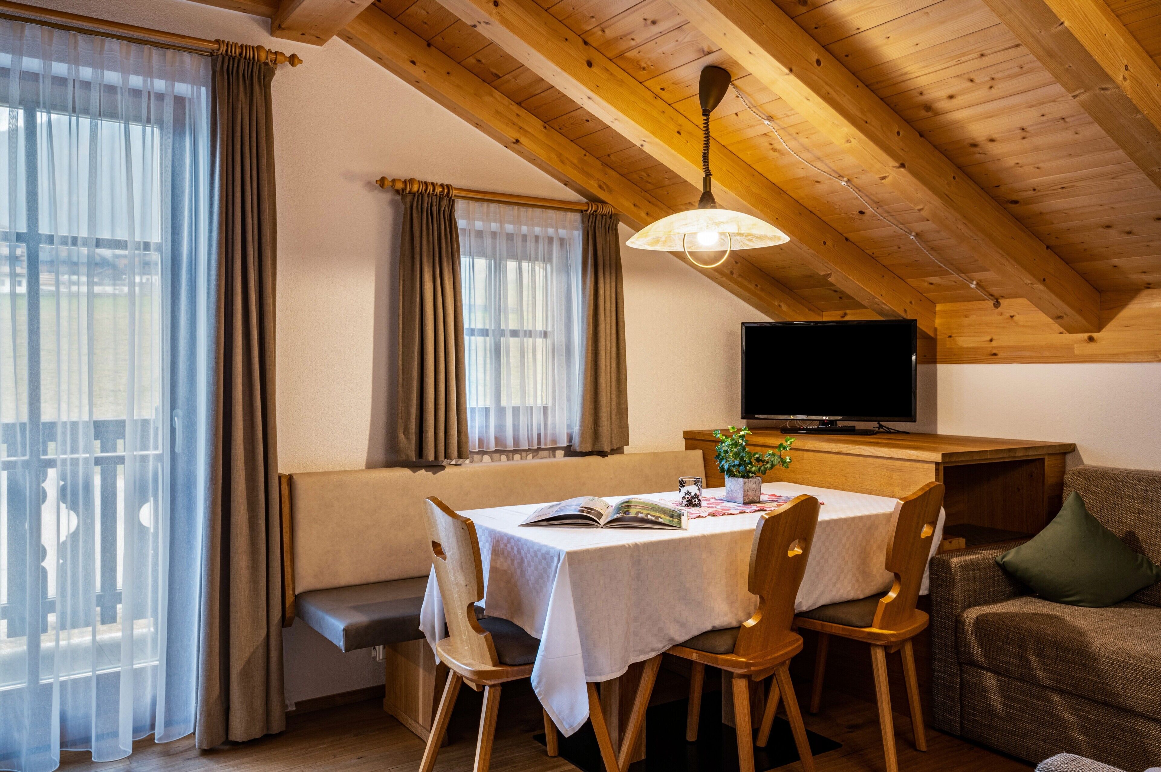 Un espace repas comprenant une table, des chaises, un banc, un téléviseur et un plafond avec des poutres en bois.