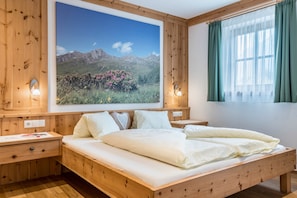 Un lit en bois avec une literie blanche, une table de chevet avec une lampe, un tableau représentant un paysage de montagne accroché au mur, et une fenêtre avec des rideaux verts.