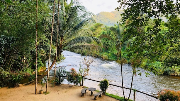 Vista a la orilla del río con un puente de madera, zona arenosa con una mesa y bancos, y abundante vegetación.
