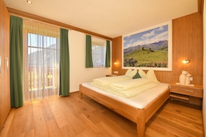 Un dormitorio con cama de madera, cortinas verdes, un ventanal con vistas a montañas y una lámpara de pared.