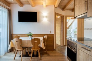A dining area with a table and chairs, a flat-screen TV mounted on the wall, wooden cabinets, and a kitchen area with a stove and oven.