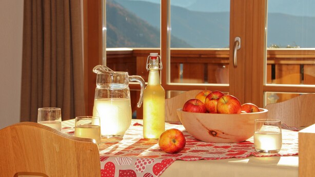 Ein Tisch mit einer Karaffe, einer Flasche, einer Obstschale und Gläsern, vor einem Fenster mit Blick auf die Berge.