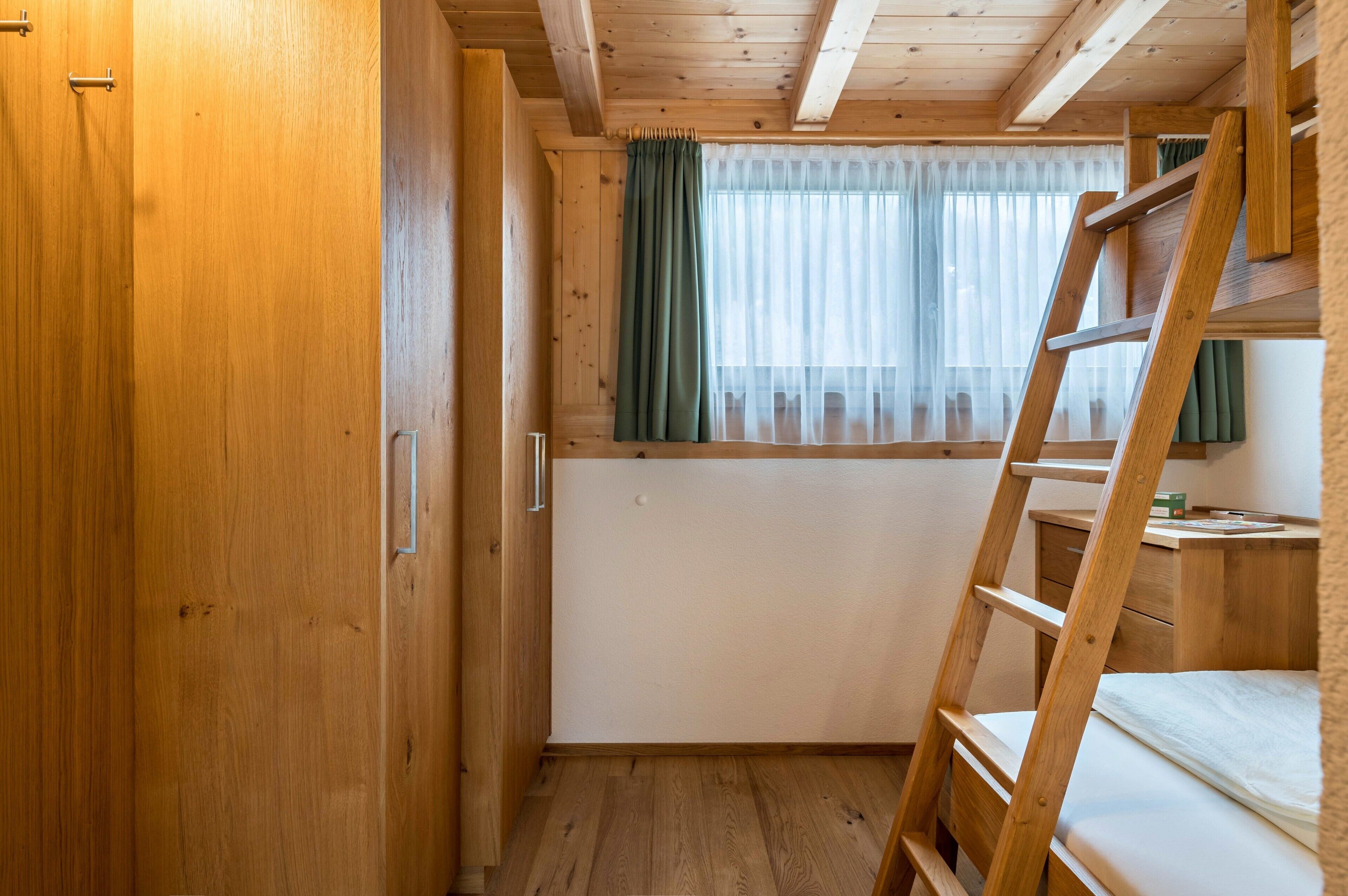 A wooden interior with a bunk bed, a ladder, a window with curtains, and a radiator.