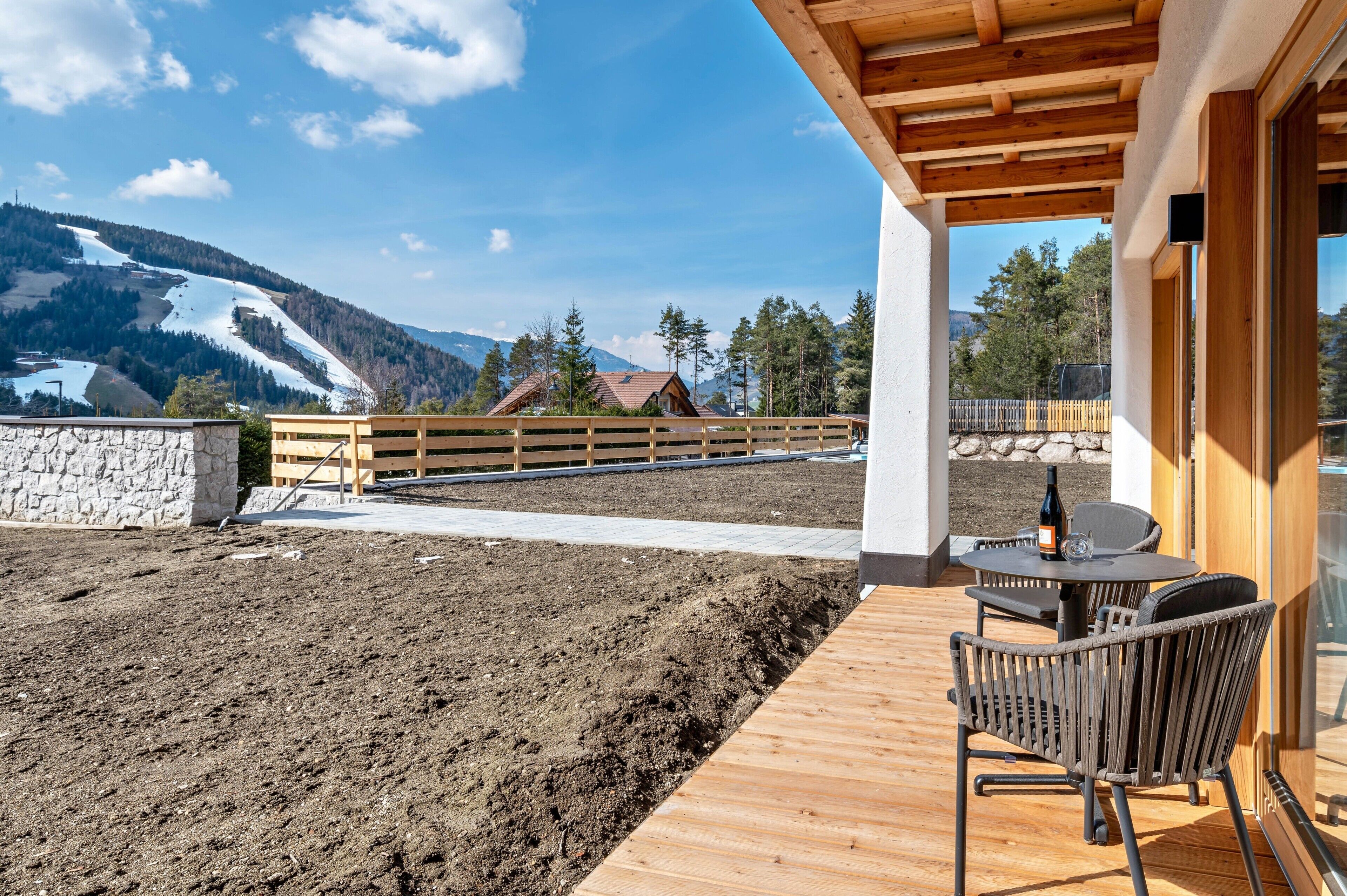 Eine Holzterrasse mit Tisch und Stühlen, eine Steinmauer und Blick auf Berge und Häuser.