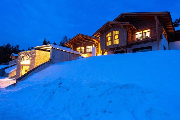A wooden chalet with a stone entrance, a balcony, and a snow-covered slope.