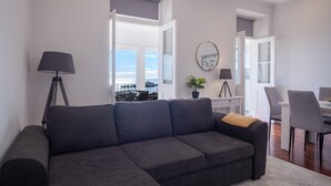 A modern living room with a grey sofa, a floor lamp, a dining area with a white table and chairs, and a large window with a view.