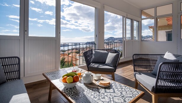 A balcony with a view of the city, a wicker chair, a sofa, and a table with fruits and a teapot.